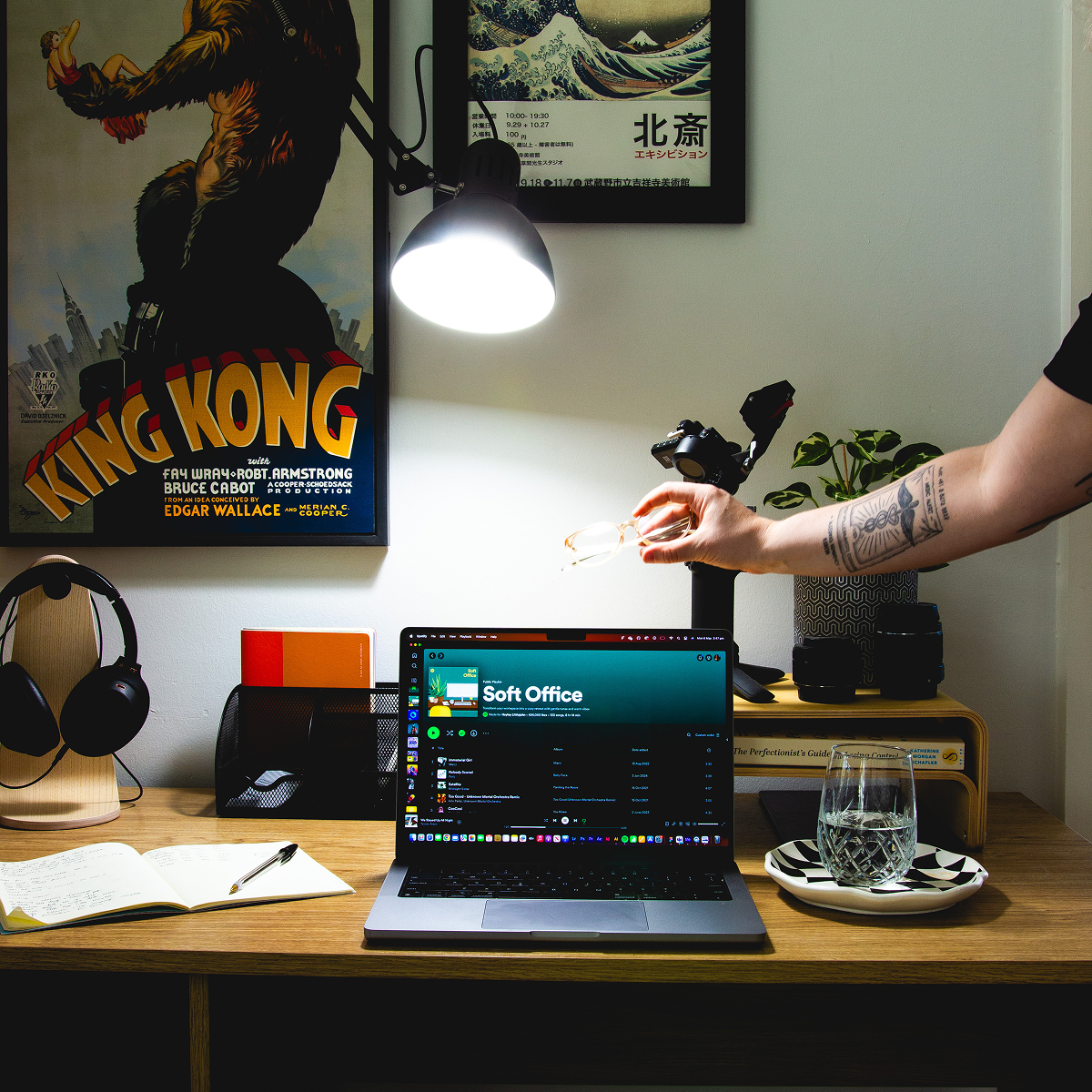 A laptop on a wooden desk displaying a software interface, with a person's tattooed arm reaching across from the right side. The desk is styled with a glass of water, an open notebook, headphones, and a retro radio, set against a wall decorated with vintage posters under a glowing desk lamp. Powered by a LIFX SuperColor 1600lm A21 smart light.