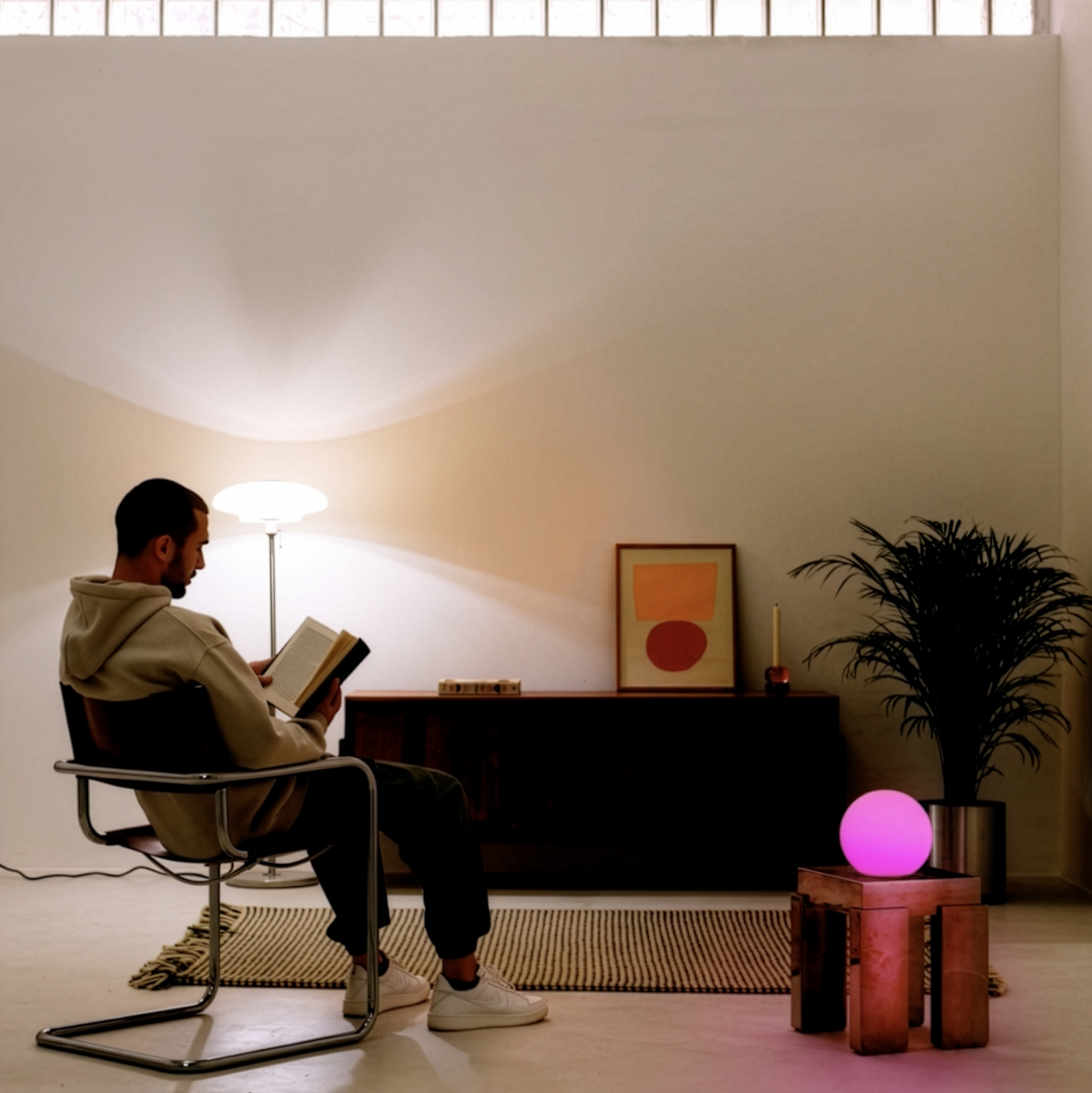 A man sits in a modern chair reading a book in a minimalist, warmly lit room. A glowing pink orb lamp sits on a small wooden table next to a potted plant, while a floor lamp illuminates the wall behind him. Powered by a LIFX SuperColor 1600lm A21 smart bulb. 